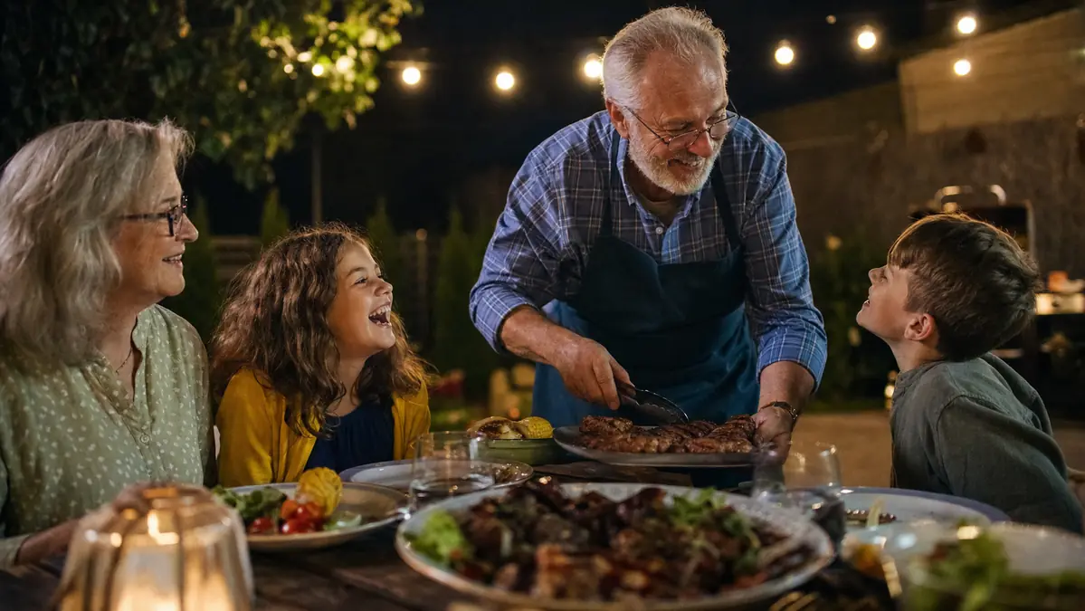 Carl, his wife Bonnie, and grandkids Mia and Owen sitting around the patio table at night while burgers finish on the lit-up grill in the background