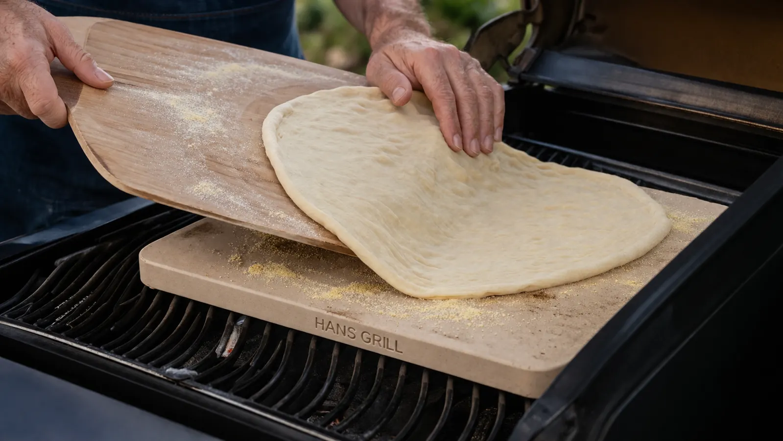 A hand in an oven mitt sliding a raw pizza off a wooden peel onto a preheated stone on the grill