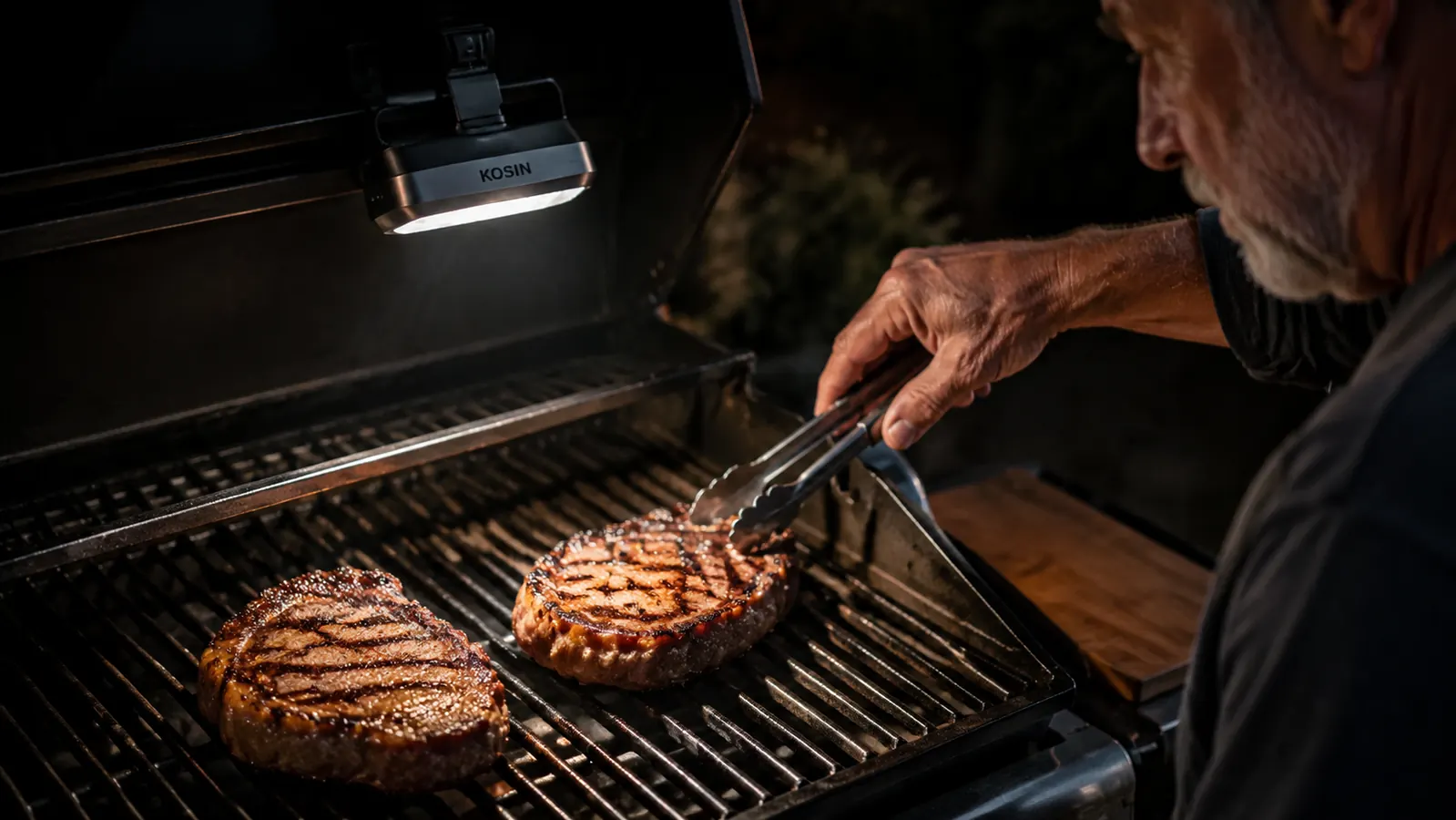 Two ribeye steaks on a grill grate clearly lit by an overhead LED light, grill marks and juices visible