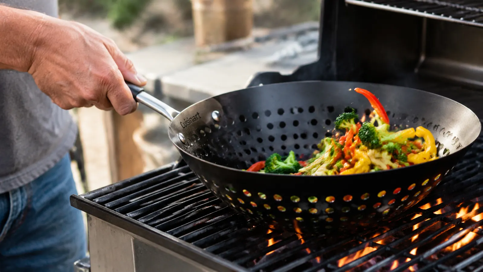 Close-up of hands tossing sliced peppers, onions, and zucchini in a perforated stainless grill wok over open flame