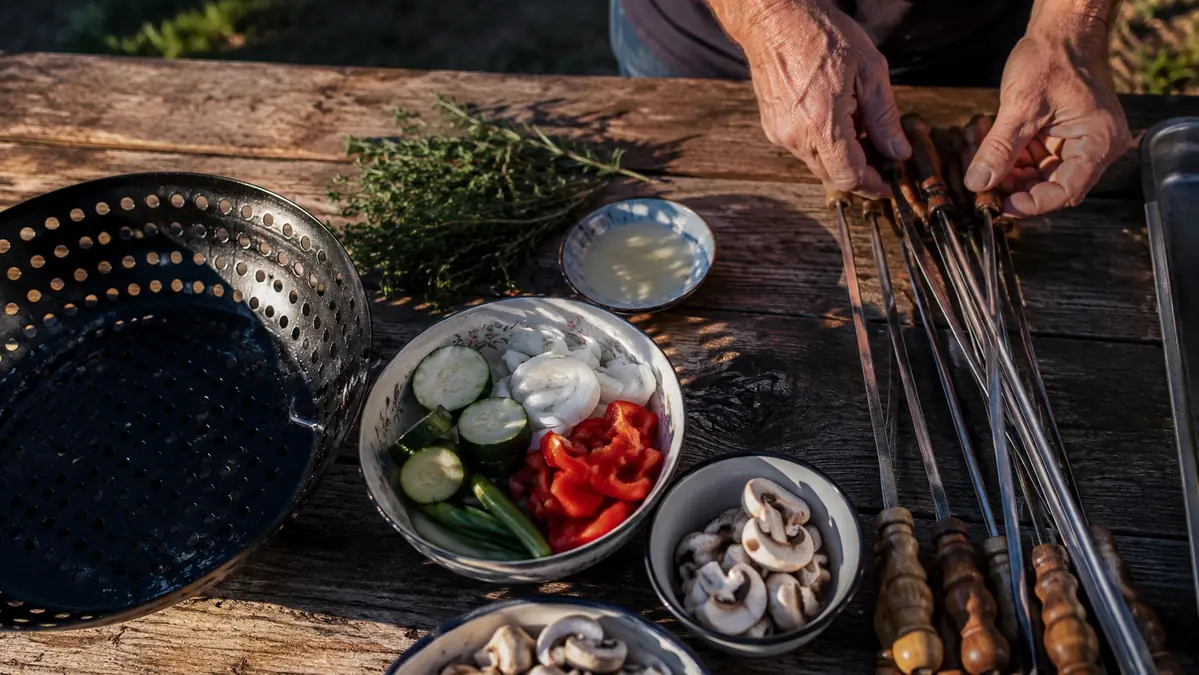 A perforated grill wok basket and a set of metal skewers laid side by side on a picnic table next to fresh cut peppers, zucchini, and onions