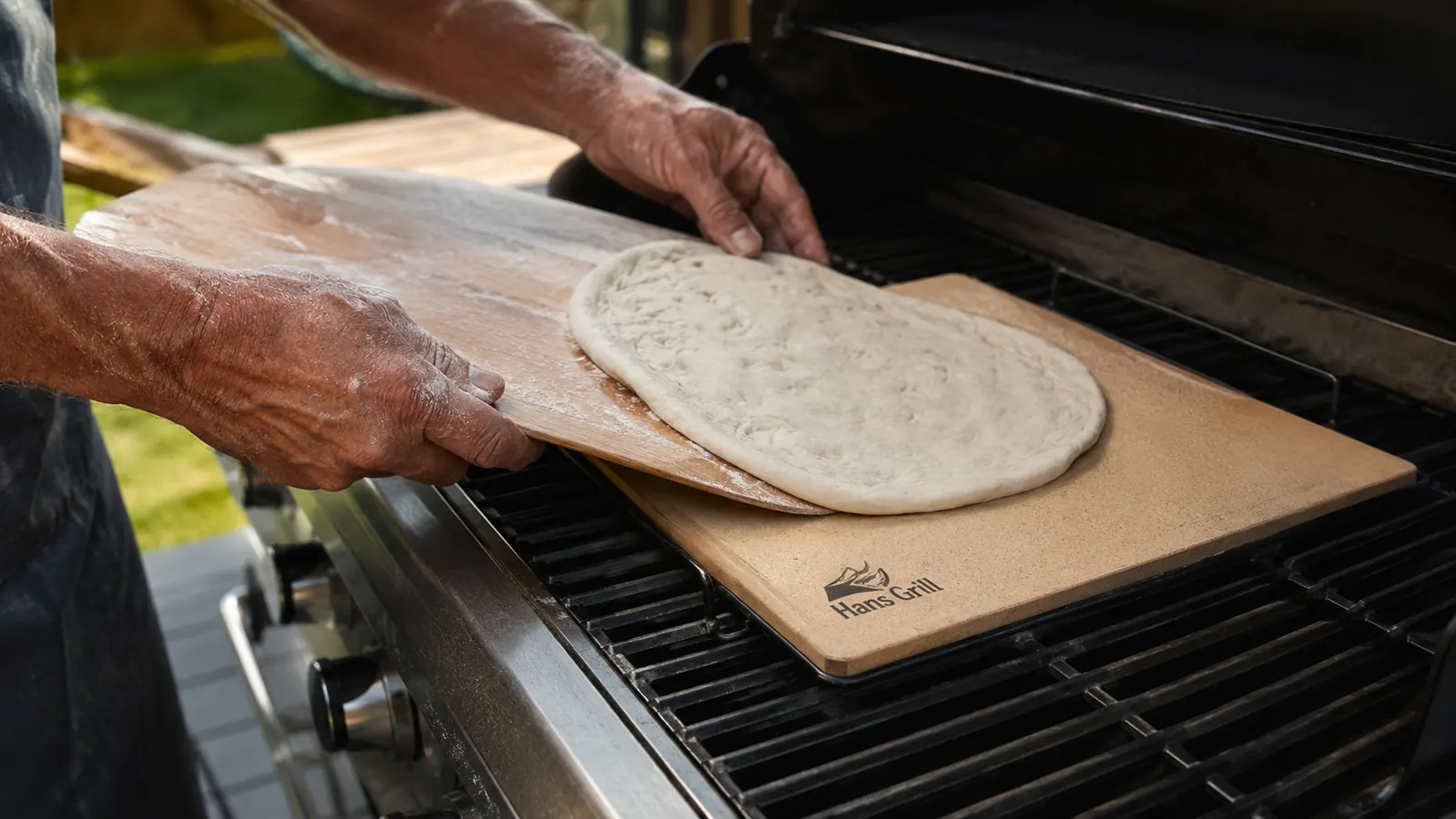 Close-up of hands sliding a raw pizza off a wooden peel onto a preheated stone on the grill