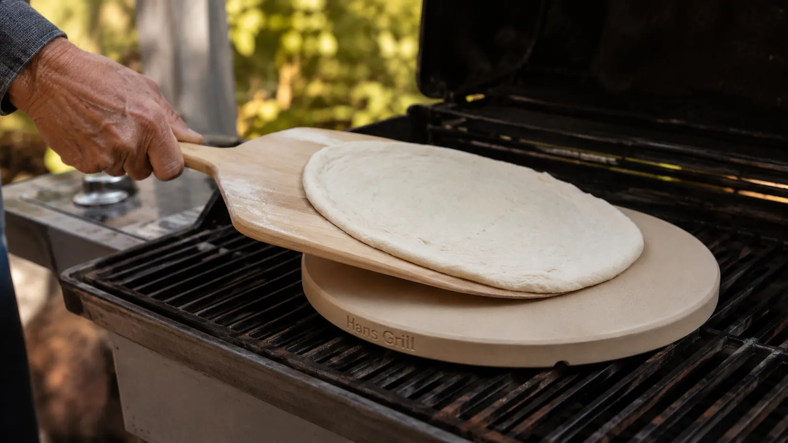 Close-up of a hand sliding a pizza off a pizza peel onto the Hans Grill cordierite stone already heating on the grill grates